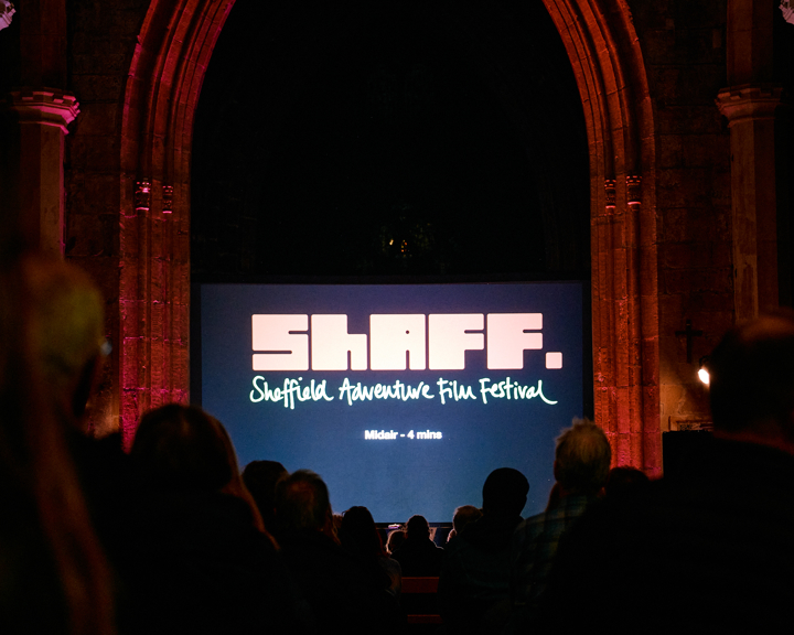 A large cinema screen, with the Sheffield Adventure Film Festival logo displayed on it, bathes an audience in a low light in Sheffield Cathedral.