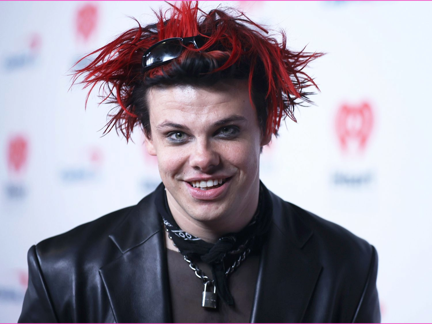 Person wearing a black leather jacket and layered necklaces, including a padlock pendant, with a black scarf tied around the neck. Hair is styled in spiky red and black strands with sunglasses resting on top. The background features a white backdrop with red heart-shaped logos.