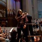 A singer stands at the front of an orchestra, holding a microphone during a rehearsal or performance in a grand concert hall. Musicians with string instruments sit in the foreground, while a conductor stands on a podium nearby. Large organ pipes and ornate architectural details form the backdrop, illuminated by warm stage lighting.