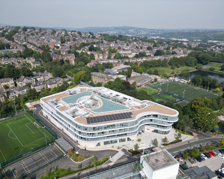 Aerial view of a modern, curved glass building with rooftop solar panels and a central circular feature, surrounded by sports fields and parking areas. In the background, a residential neighborhood with rows of houses and green spaces stretches across rolling hills under a hazy sky.