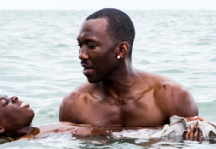 Two people are in shallow seawater near a shoreline. One person supports another, who is floating on their back with their head above the water. Gentle waves ripple around them, and open water stretches into the background. The scene appears to depict a water rescue, lifesaving exercise or dramatic moment taking place at sea.