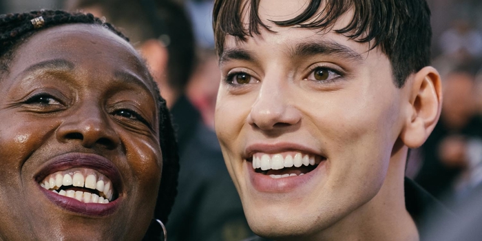 Two people standing close together in an outdoor setting, with a crowd and buildings visible in the background. The image shows bright stage lights overhead and blurred architectural details behind them, suggesting an event or gathering in an urban area. One person is wearing a dark outfit, while the other has braided hair and a patterned top.