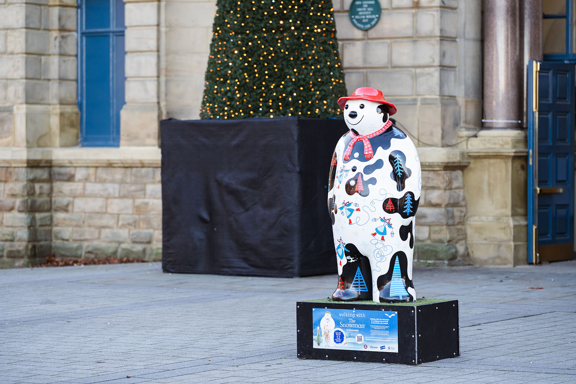 A decorated bear sculpture wearing a red hat and scarf stands on a black platform in front of a stone building. The bear is painted with colorful patterns, including trees, snowflakes, and abstract designs. Behind it, a large Christmas tree wrapped in lights is partially visible, and the setting appears to be an outdoor festive display.