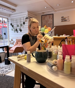 A cozy art studio with a person seated at a table painting a ceramic item. The table is covered with paint bottles, brushes, and a mug. In the background, there are shelves with decorative items, framed artwork on the wall, and a window displaying a street view with a red shop sign. The space is decorated with plants and has warm lighting.