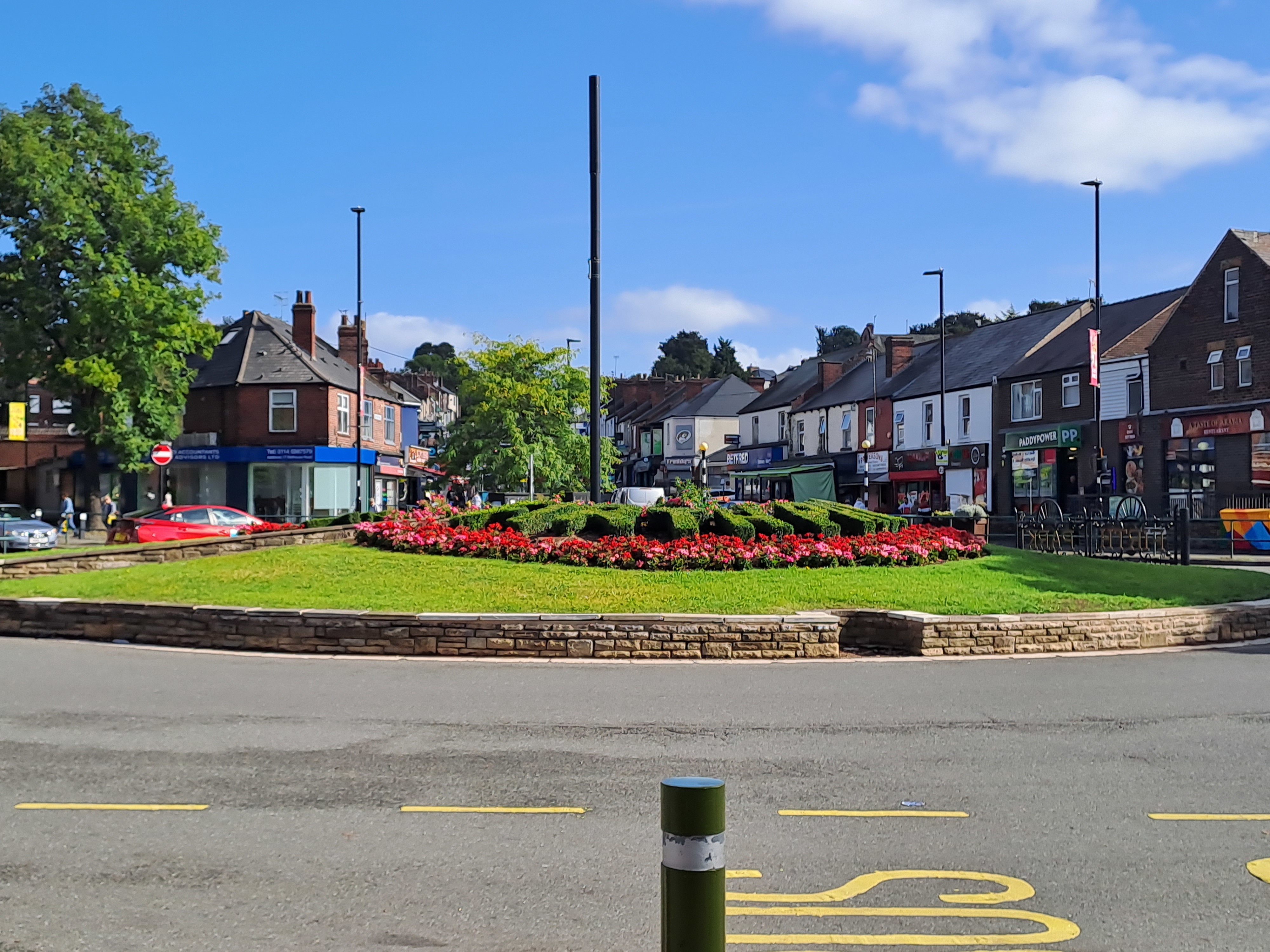 own center roundabout with a neatly maintained circular flower bed featuring red blooms and green shrubs, surrounded by a low stone wall. Shops and businesses line the street in the background, including a pharmacy and other storefronts, under a bright blue sky with scattered clouds.