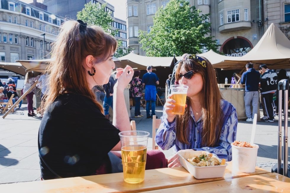 Two women in sunglasses sit drinking a pint of beer with food stall in the background 