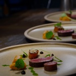 Close-up of a plated fine-dining dish featuring slices of seared meat, cylindrical cuts, bright orange vegetables, green herb leaves, and artistic drizzles of sauce on a neutral-toned plate.