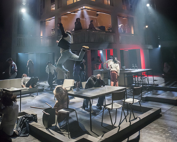 A dramatic stage performance featuring multiple actors in an industrial-style set. The foreground shows scattered tables and chairs with performers in dynamic poses, including one person leaping into the air. The background includes a two-level structure with additional actors visible on the upper floor, illuminated by theatrical lighting in shades of blue and red. The scene conveys high energy and movement.