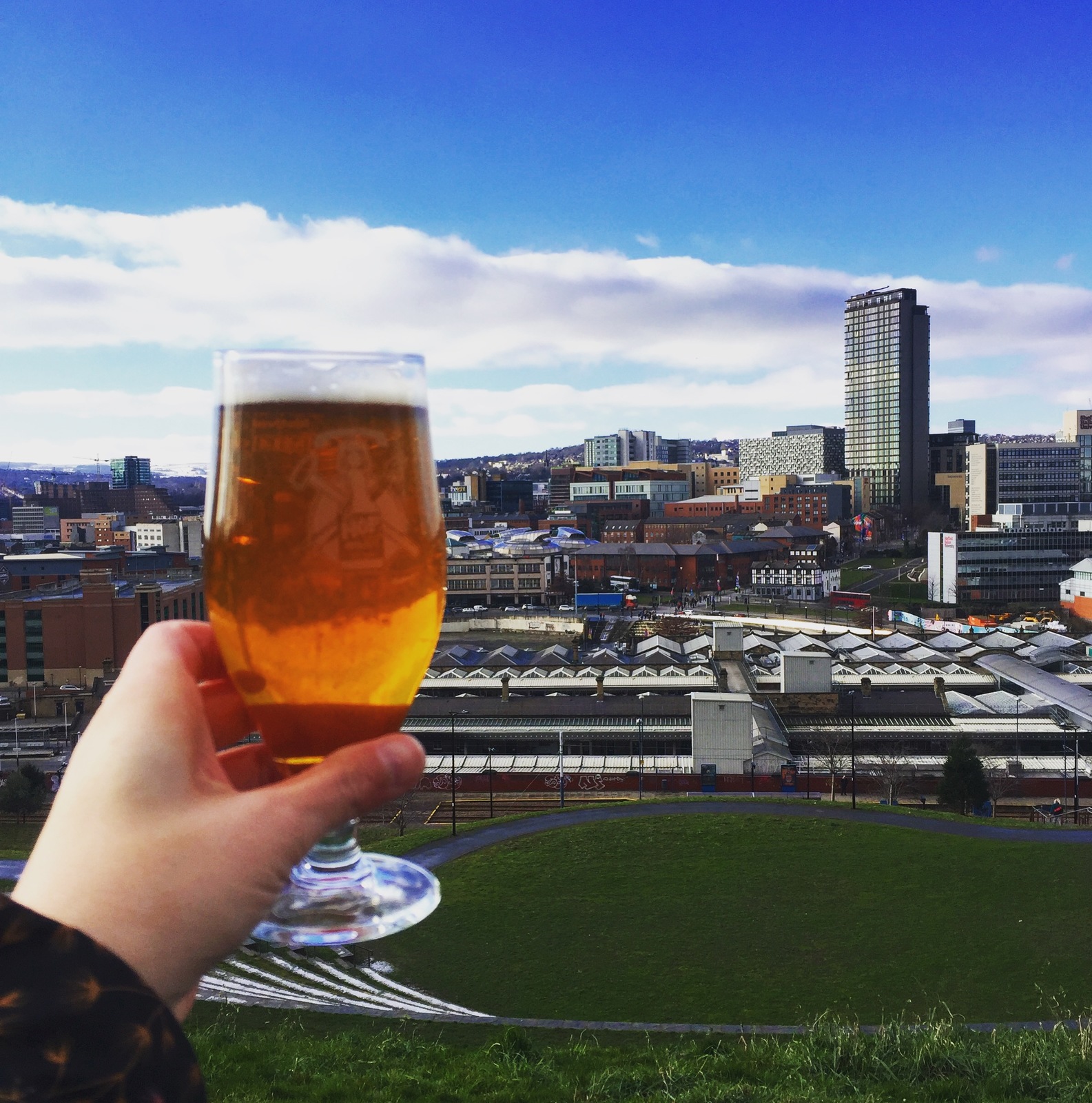 A hand holding a glass of beer in front of the Sheffield skyline on a sunny day.