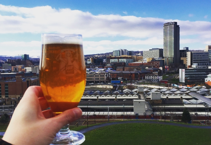 A hand holding a glass of beer in front of the Sheffield skyline on a sunny day.