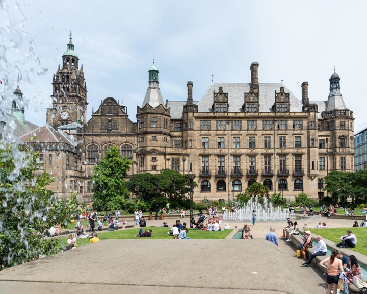 View of Sheffield Town Hall from the Peace Gardens, showing the historic stone building with ornate towers and spires. In the foreground, people are sitting and relaxing around fountains and green lawns under a bright sky.