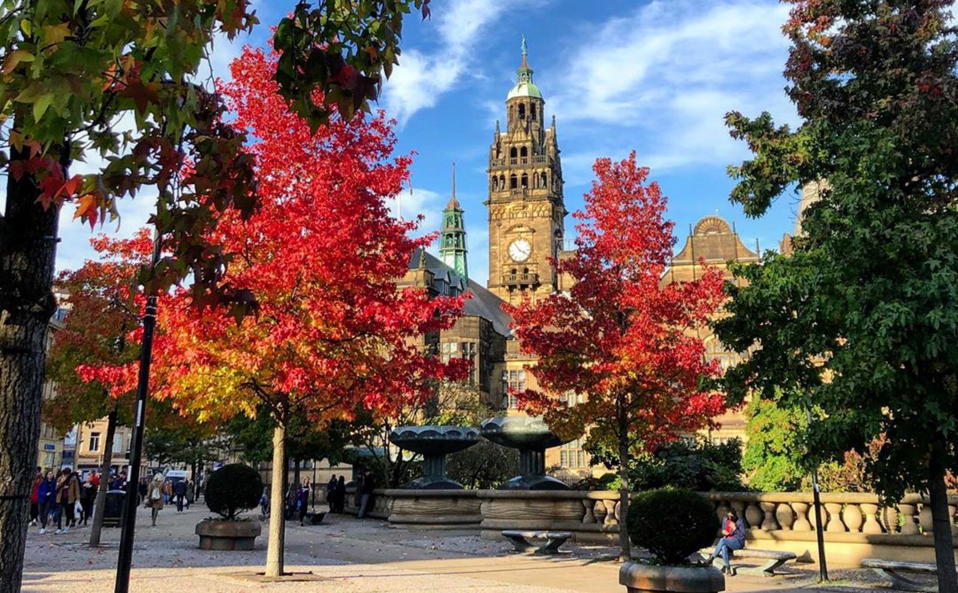 City square with vibrant autumn trees in shades of red and orange, framing a historic building with a tall clock tower in the background. The scene includes ornate fountains, stone balustrades, and people walking around under a bright blue sky with scattered clouds.