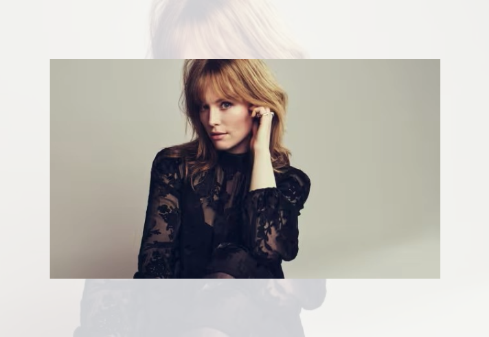 Studio portrait of a person with shoulder-length auburn hair wearing a black lace top with sheer floral-patterned sleeves. The person is seated against a plain light grey background, looking towards the camera with one hand raised to the side of the head. Soft lighting and muted tones create a polished, professional promotional-style image.