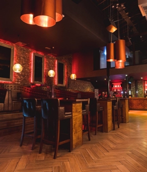 Interior shot of Crystal. Modern bar interior with warm lighting, exposed brick walls, and wooden herringbone flooring. The space features high tables with leather chairs, booth seating along the wall, and a long bar counter with illuminated shelves stocked with bottles. Copper pendant lights hang from the ceiling, adding to the stylish atmosphere.
