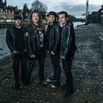 Four people dressed in dark clothing and leather jackets stand on a cobbled riverside quay beside a waterway lined with historic brick buildings. One person wears a flat cap, another has long dreadlocks, and a third wears a bowler-style hat. The overcast sky and wet cobbles create a moody urban atmosphere, with a bridge visible in the distance across the river.