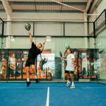 Two players on an indoor padel court, one jumping to hit the ball mid‑air while the other gets into position, with glass walls and vibrant sports-themed graphics in the background