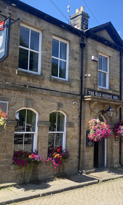 The Old Horns Inn photographed on a sunny day, showcasing its traditional stone façade, arched windows, colourful hanging baskets and cobbled street setting.