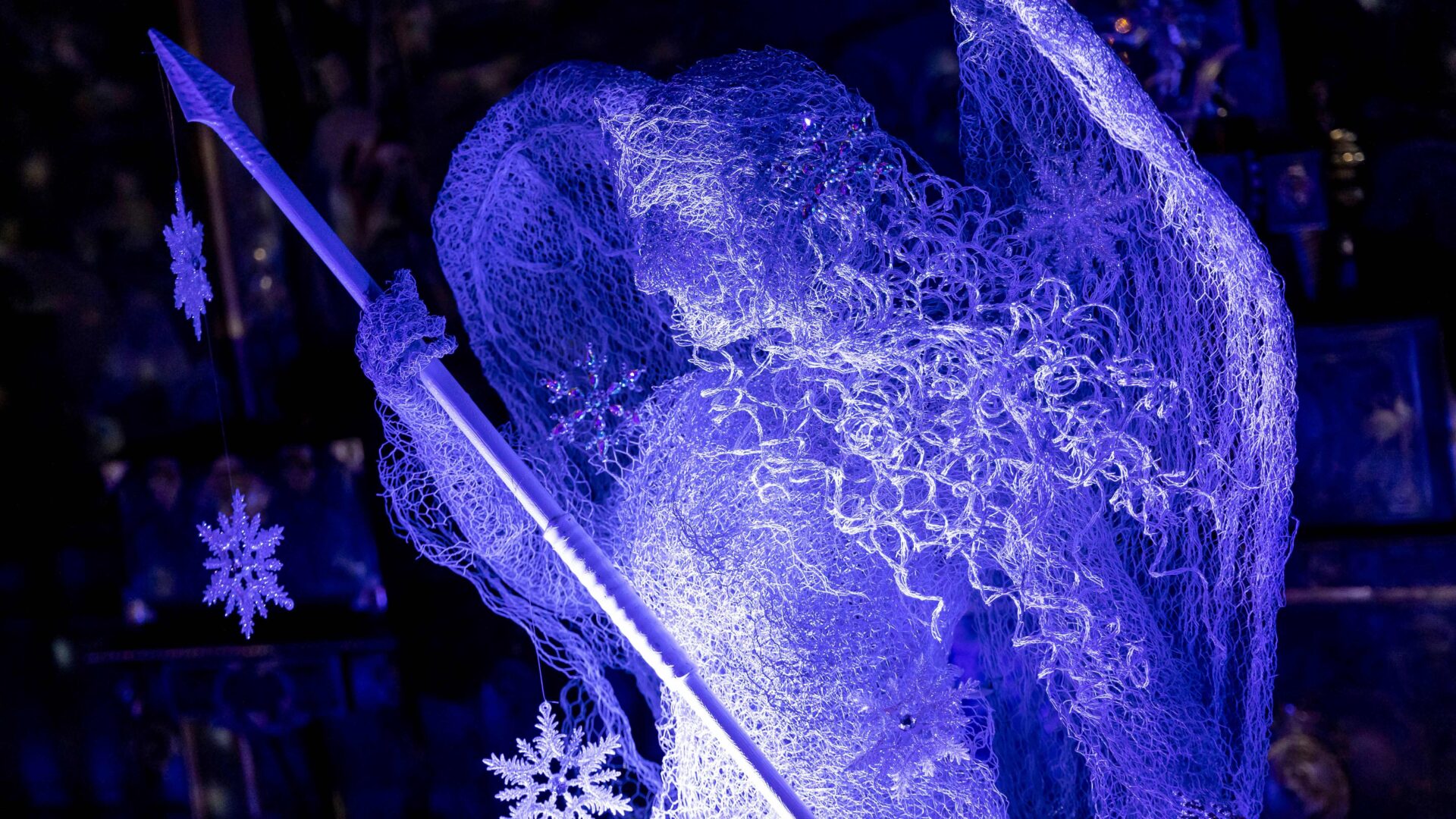 A striking sculpture of an angel made from intricate wire mesh, illuminated in vivid blue light. The angel holds a long spear-like staff and has large, detailed wings. Snowflake ornaments hang nearby, adding to the wintry, festive atmosphere. The background is dark, making the glowing figure stand out dramatically.