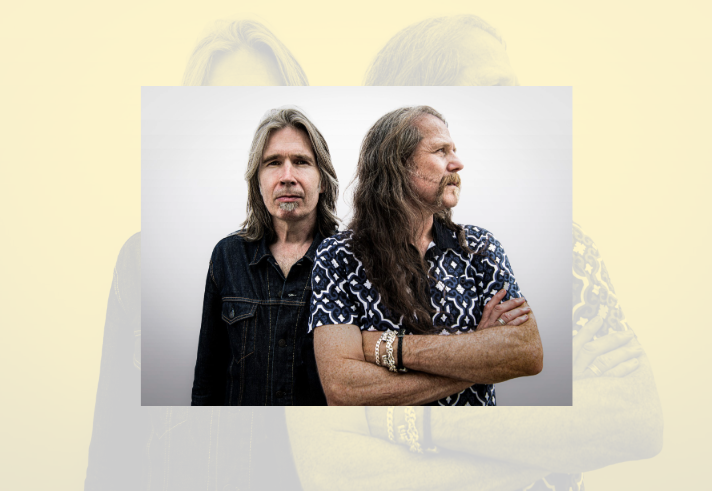 Two long-haired musicians pose against a plain light background. One stands facing the camera wearing a dark denim jacket, while the other stands slightly in front with arms folded, looking to the side and wearing a patterned short-sleeved shirt. The close-up portrait focuses on their contrasting poses and hairstyles, creating a simple, studio-style promotional image.