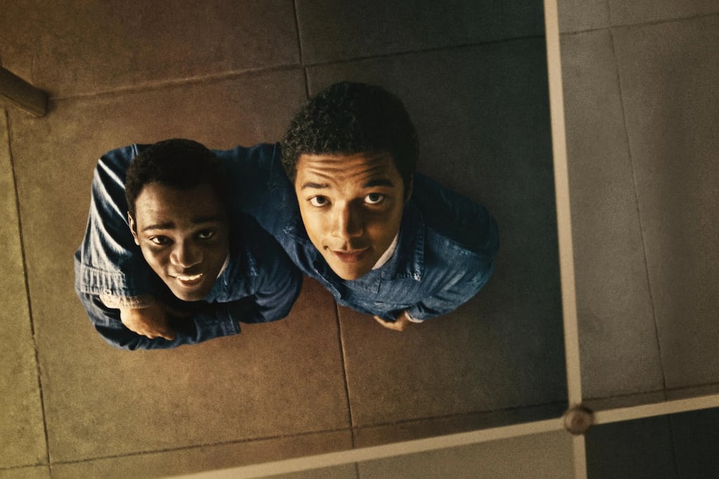 Two people wearing denim shirts are standing close together on a tiled floor, viewed from above. Their arms are positioned near their sides, and the scene is framed by a grid-like structure in the foreground, possibly part of a ceiling or partition. The lighting is warm and soft, casting subtle shadows across the tiles.