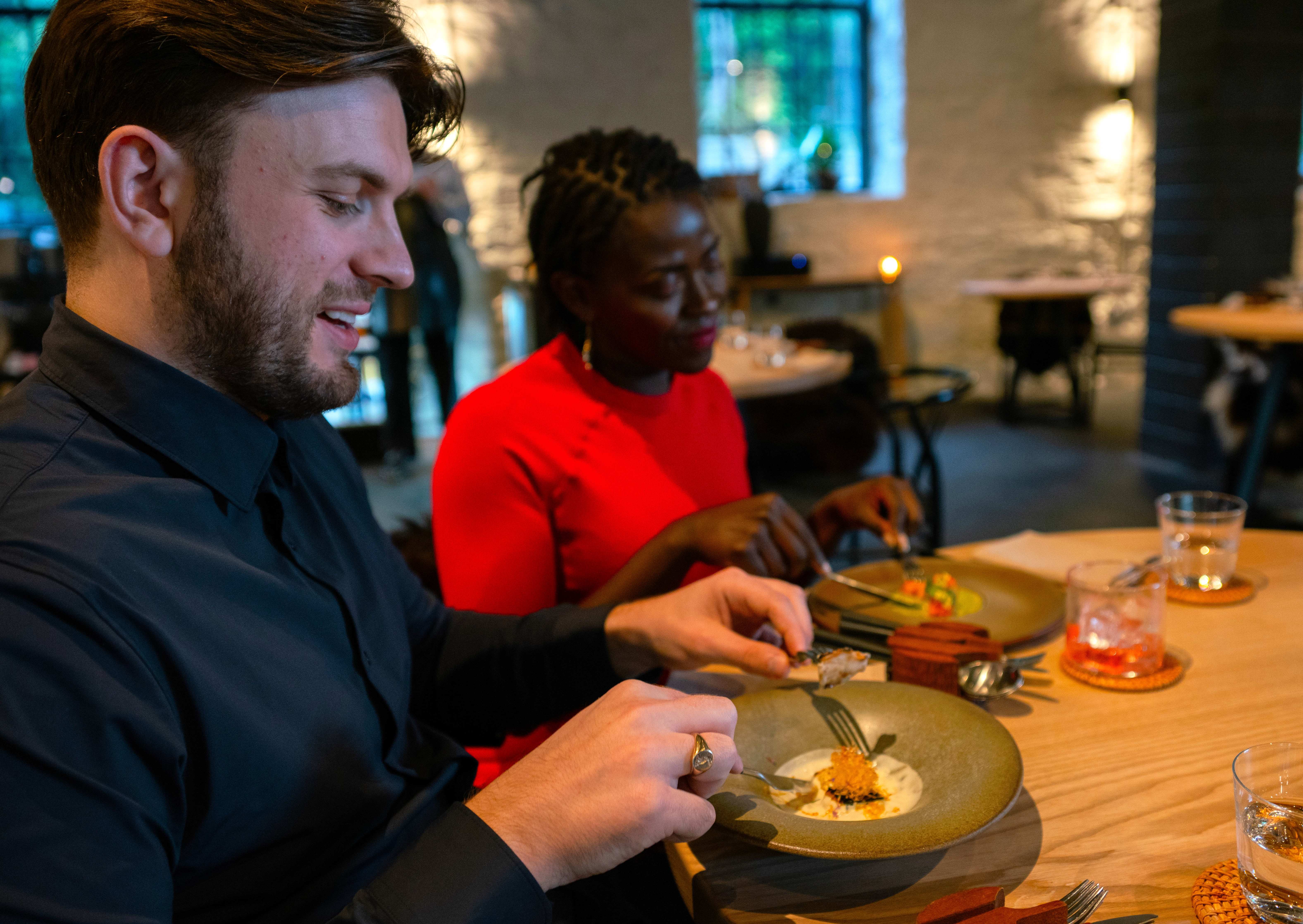 Two diners seated at a wooden table in a softly lit restaurant, eating a carefully plated dish with drinks beside them.