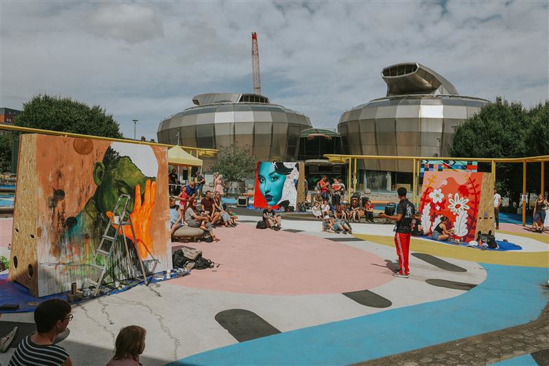 Outdoor art event in a colorful plaza featuring large painted panels with vibrant designs, including a portrait and abstract floral artwork. People are sitting and standing around the area, watching artists work. In the background, two modern, metallic, dome-shaped buildings are visible under a partly cloudy sky.
