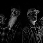 Black-and-white portrait of four people standing closely together against a dark brick wall. Dramatic side lighting illuminates their faces and clothing while much of the background remains in shadow. One person has a long white beard and a knit hat, another wears a baseball cap and jacket, and the others wear casual outdoor clothing. The high-contrast monochrome style creates a striking, atmospheric group portrait.
