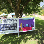A large banner displayed outdoors under a tree reads “Graysons Solicitors welcomes you to Art in the Gardens” with the website www.graysons.co.uk. The banner features a colourful illustration of an art gallery with a sculpture and arched doorway. Behind it, people are seated near white tents on a grassy area, and a wide path leads through landscaped gardens with trees and shrubs in bright sunlight.