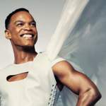 A smiling person stands confidently with hands on hips, wearing a white sleeveless outfit with a cut-out detail at the chest. Sheer white fabric billows behind them like wings, creating a dramatic effect. The background features a soft grey-blue sky with subtle sparkling highlights, giving the image a theatrical and uplifting feel.