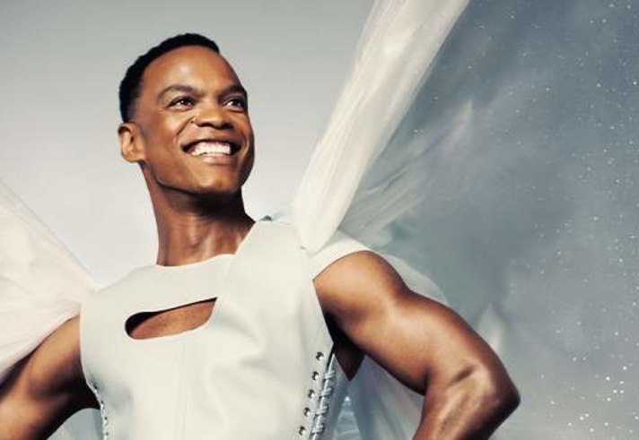 A smiling person stands confidently with hands on hips, wearing a white sleeveless outfit with a cut-out detail at the chest. Sheer white fabric billows behind them like wings, creating a dramatic effect. The background features a soft grey-blue sky with subtle sparkling highlights, giving the image a theatrical and uplifting feel.