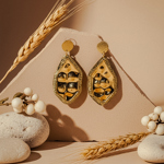 Bold handcrafted earrings with textured gold and black abstract design, displayed against a neutral backdrop with wheat stalks and smooth stones for a natural aesthetic