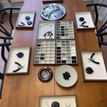 Overhead view of a jewellery-making workstation arranged on a long wooden table. Shallow trays hold pliers, scissors, measuring tapes, beads and jewellery findings sorted by colour and size. A partially assembled beaded necklace and loose black beads sit in the centre beside compartment trays filled with metal components. Black chairs surround the table, suggesting a craft workshop or handmade jewellery production session in progress.