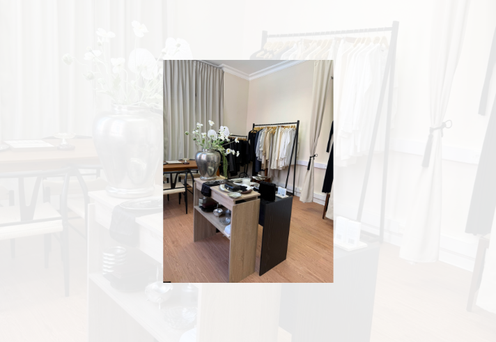 Interior of a minimalist boutique or showroom with cream walls, tall curtains and wooden flooring. In the foreground, a display table holds a large silver vase filled with white flowers alongside trays, dishes and accessories. Behind it, two clothing rails display neatly arranged garments in neutral shades including white, beige, grey and black. A wooden table and chairs sit to the left, creating a calm, curated retail setting.