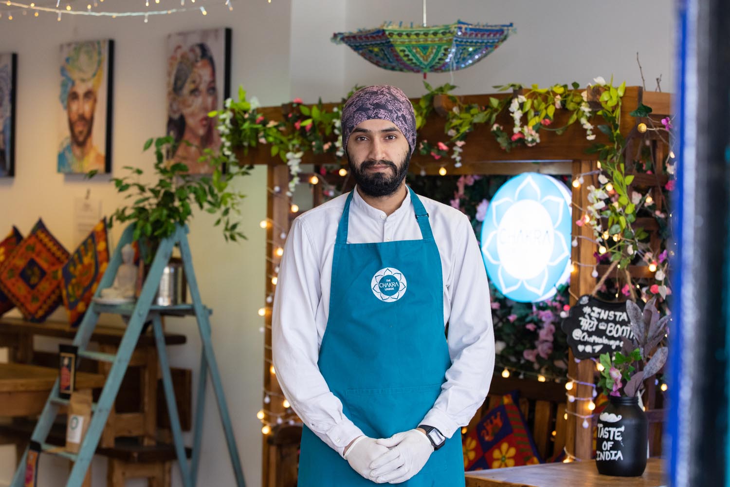 A person wearing a teal apron and white gloves stands inside a decorated shop or café. The background features colorful patterned cushions, hanging string lights, green plants, and a wooden display with flowers. A round illuminated sign and a chalkboard reading “Taste of India” are visible behind the person.