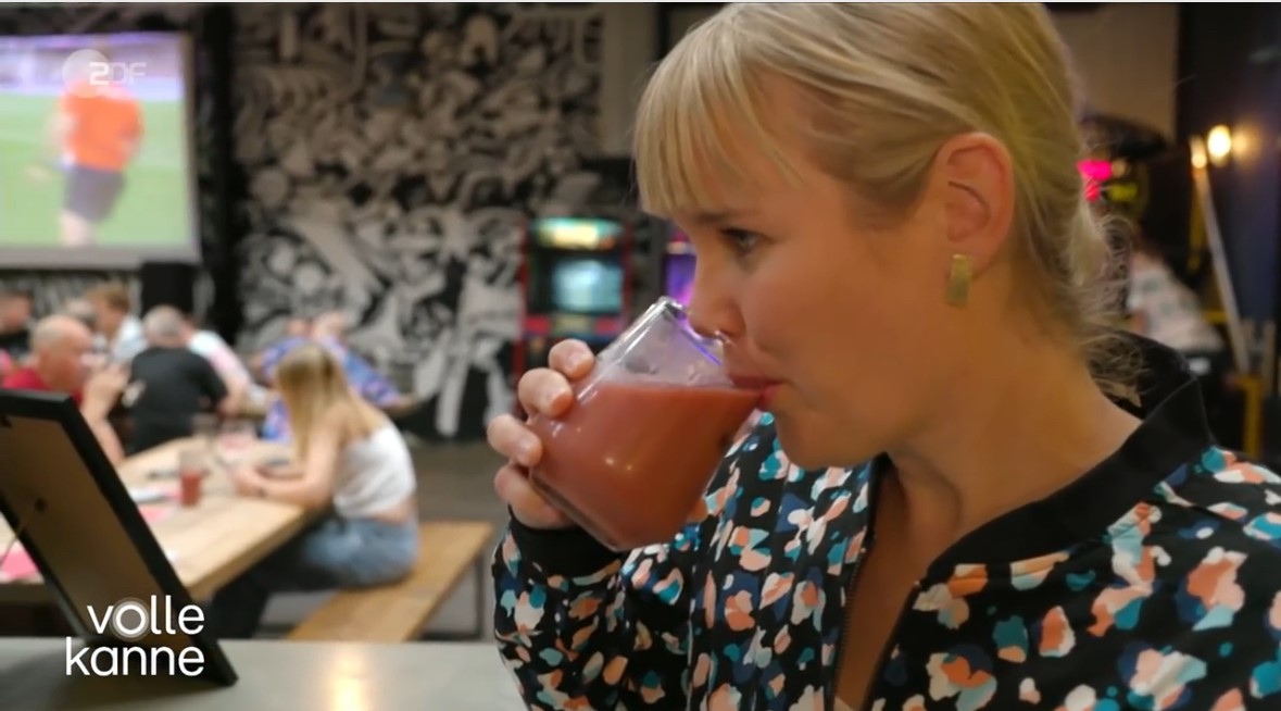 “Person holding a glass of pink beverage inside a casual dining area with wooden tables and benches. Several people are seated in the background, and a large screen showing a sports game is visible on the wall along with black-and-white mural artwork. Text on the bottom left reads ‘volle kanne.’”