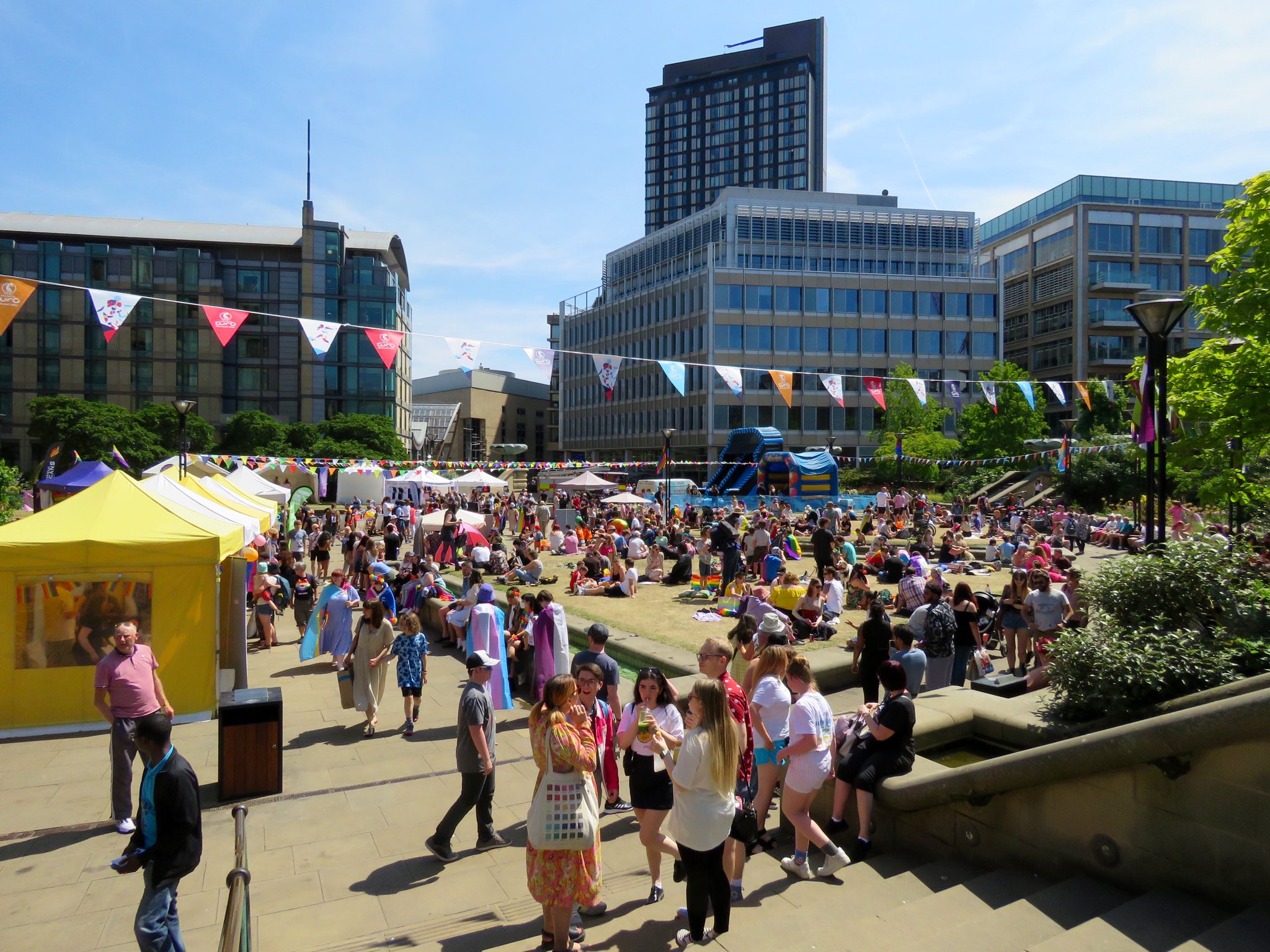 Outdoor event in a city square on a sunny day, with crowds gathered on grass and paved areas. Colorful bunting is strung overhead, and yellow and white tents line the left side. People are sitting on picnic blankets and walking around, with modern office buildings and a tall tower in the background.
