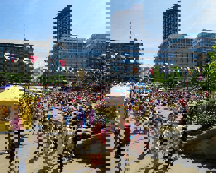 Outdoor event in a city square on a sunny day, with crowds gathered on grass and paved areas. Colorful bunting is strung overhead, and yellow and white tents line the left side. People are sitting on picnic blankets and walking around, with modern office buildings and a tall tower in the background.