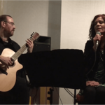 A live music performance in an indoor venue. One performer sits playing an acoustic guitar while another sits beside them holding a microphone and singing. A music stand is positioned between the performers, with a speaker visible in the background. The intimate setting is softly lit, focusing attention on the acoustic duo and their performance.