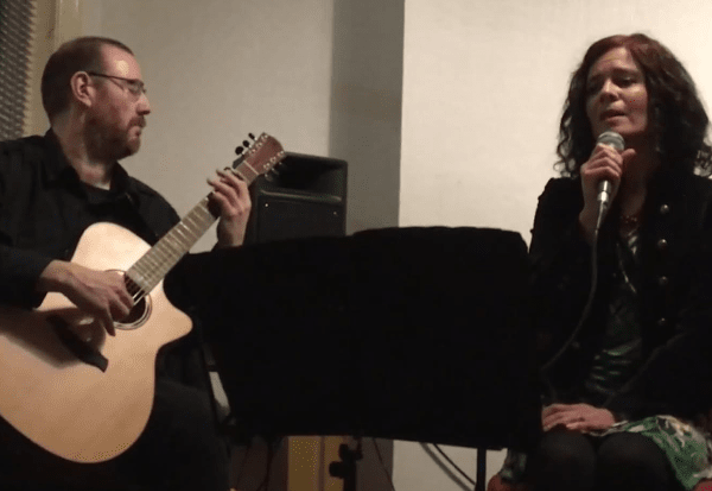 A live music performance in an indoor venue. One performer sits playing an acoustic guitar while another sits beside them holding a microphone and singing. A music stand is positioned between the performers, with a speaker visible in the background. The intimate setting is softly lit, focusing attention on the acoustic duo and their performance.