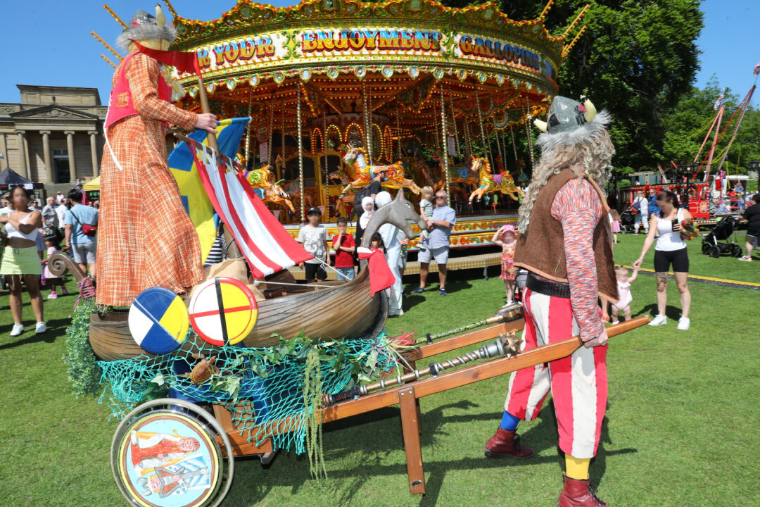 Two performers in Viking-themed costumes stand beside a decorated cart shaped like a wooden boat, complete with shields, a striped sail, and fishing nets. The cart is on grass in front of a brightly lit carousel with colourful horses. People in summer clothing watch the scene, and trees and a historic building are visible in the background, creating a lively festival atmosphere.
