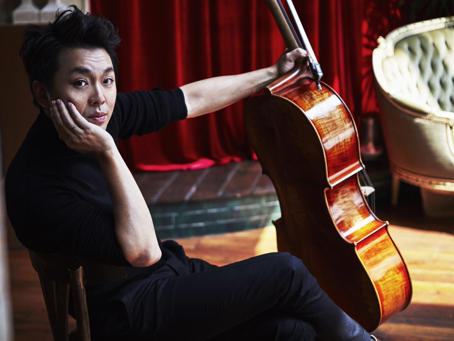 A person dressed in black is seated on a wooden chair indoors, holding a cello upright by its neck. The cello has a polished wooden finish and rests on the floor beside the chair. The background features a red curtain and a light-coloured upholstered chair, with warm wooden flooring adding to the intimate setting.