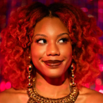 Close-up portrait of a smiling woman with vibrant curly red hair, looking to the side. She wears dramatic eye make-up, dark lipstick, large dangling earrings and an ornate gold necklace. Warm red and pink lighting illuminates her face, while a sparkling, out-of-focus backdrop creates a glamorous and festive atmosphere.
