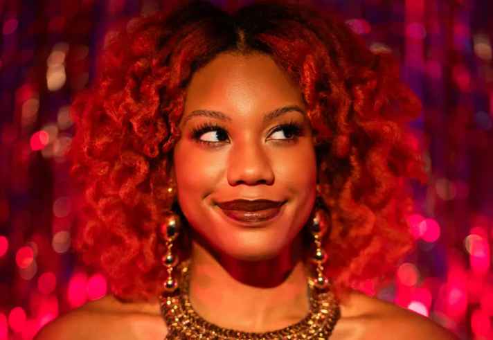 Close-up portrait of a smiling woman with vibrant curly red hair, looking to the side. She wears dramatic eye make-up, dark lipstick, large dangling earrings and an ornate gold necklace. Warm red and pink lighting illuminates her face, while a sparkling, out-of-focus backdrop creates a glamorous and festive atmosphere.