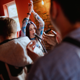 Indoor scene with exposed brick wall and warm lighting. A person stands in front of a microphone with arms raised, wearing a patterned long-sleeve top. Several people are gathered around, partially visible in the foreground, creating a lively atmosphere.