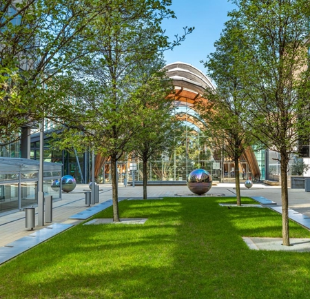 A landscaped courtyard with a row of evenly spaced trees on a bright green lawn, surrounded by modern glass and stone buildings. In the center, reflective silver spheres are placed along the walkway, leading toward a large glass atrium with an arched wooden frame. The area is clean and contemporary, with clear blue skies above.