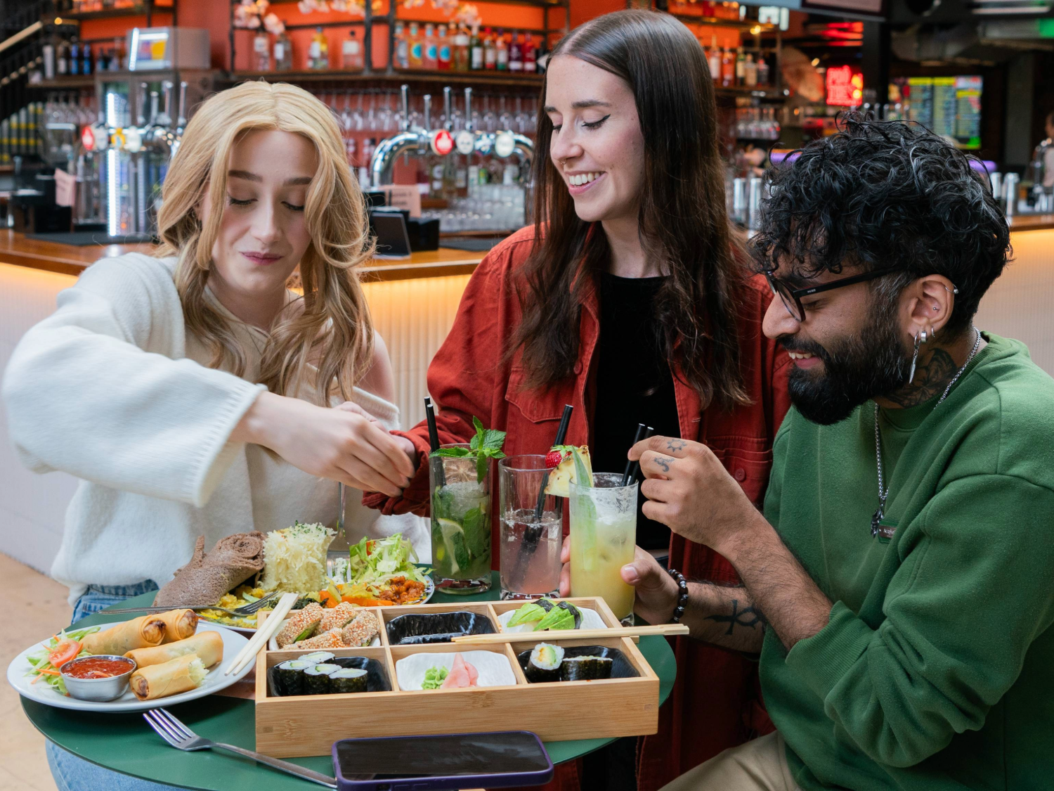 Three people seated at a round green table in a lively indoor food hall with a bar in the background. The table is filled with a wooden bento-style tray containing assorted sushi, nigiri, and pickled ginger, alongside plates of salad, bread, and spring rolls. Several tall glasses of colourful drinks with fruit and herb garnishes are also on the table. The setting is bright and vibrant, with shelves of bottles visible behind the bar.