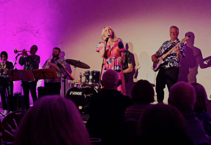 A live band performs on stage under purple lighting, featuring musicians playing trumpet, saxophone, drums, electric guitar, and bass. A vocalist stands at the centre wearing a colourful patterned dress, singing into a microphone. The audience is seated in the foreground, watching the performance against a plain wall backdrop.