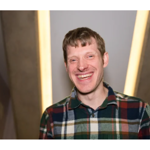 Portrait of a person standing indoors, facing the camera and wearing a multicoloured check-pattern shirt. The person is centred in the frame between two angled walls, with warm vertical light strips in the background creating a symmetrical composition. The image is sharply focused on the person, while the background is softly blurred.