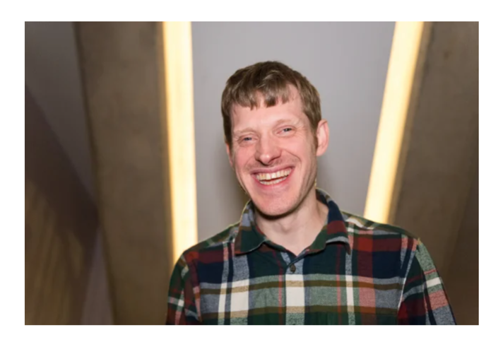 Portrait of a person standing indoors, facing the camera and wearing a multicoloured check-pattern shirt. The person is centred in the frame between two angled walls, with warm vertical light strips in the background creating a symmetrical composition. The image is sharply focused on the person, while the background is softly blurred.