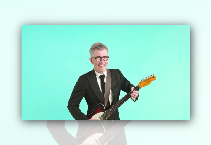 Studio portrait of a person wearing a black suit, white shirt and black tie, standing against a mint-green background while holding an electric guitar. The guitar has a dark red and black body with a light-coloured neck. The person is centred in the frame and wears round glasses, with a guitar strap over one shoulder.